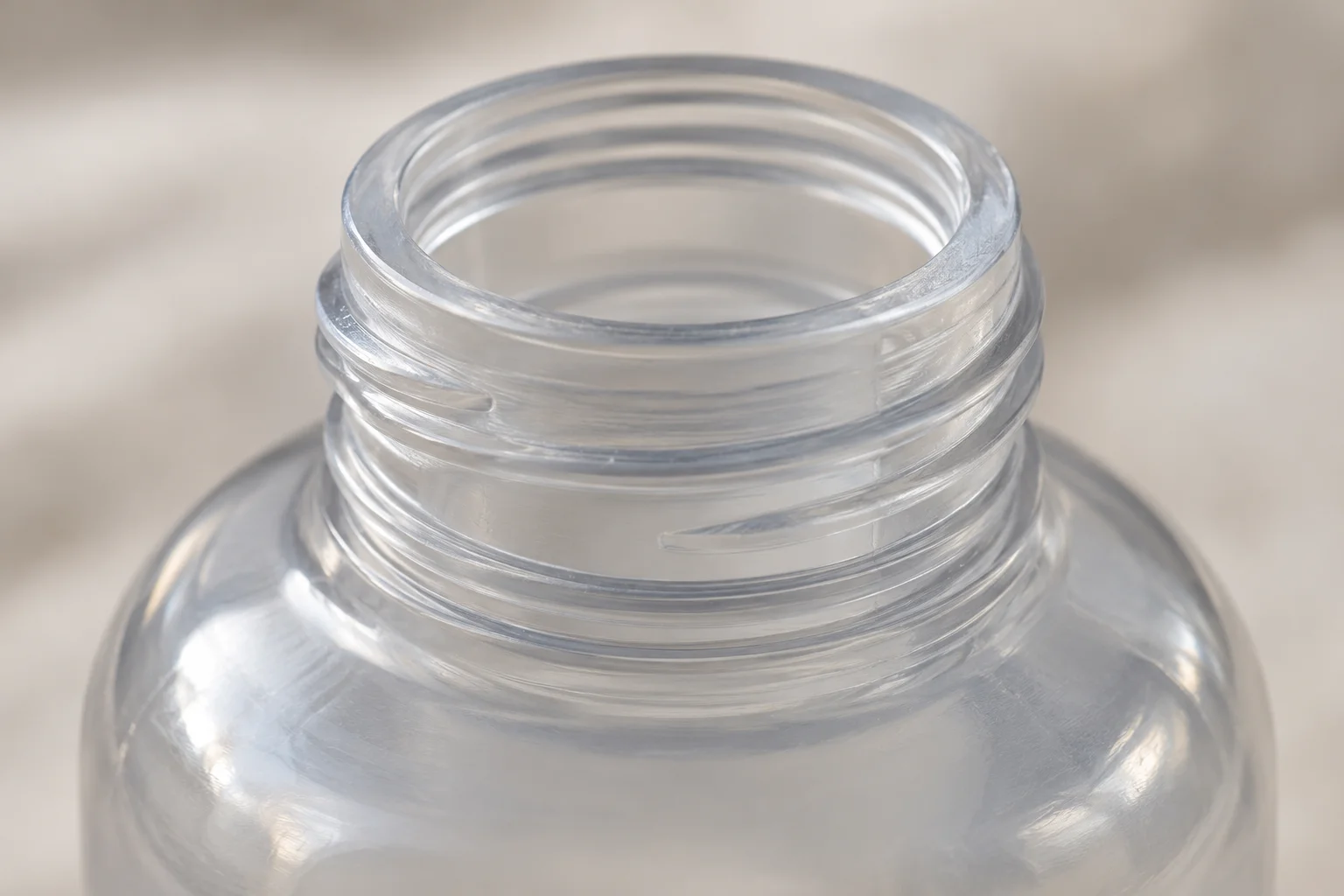 Close-up of a clear bottle's open neck, flat top rim, molded threads and rounded shoulder