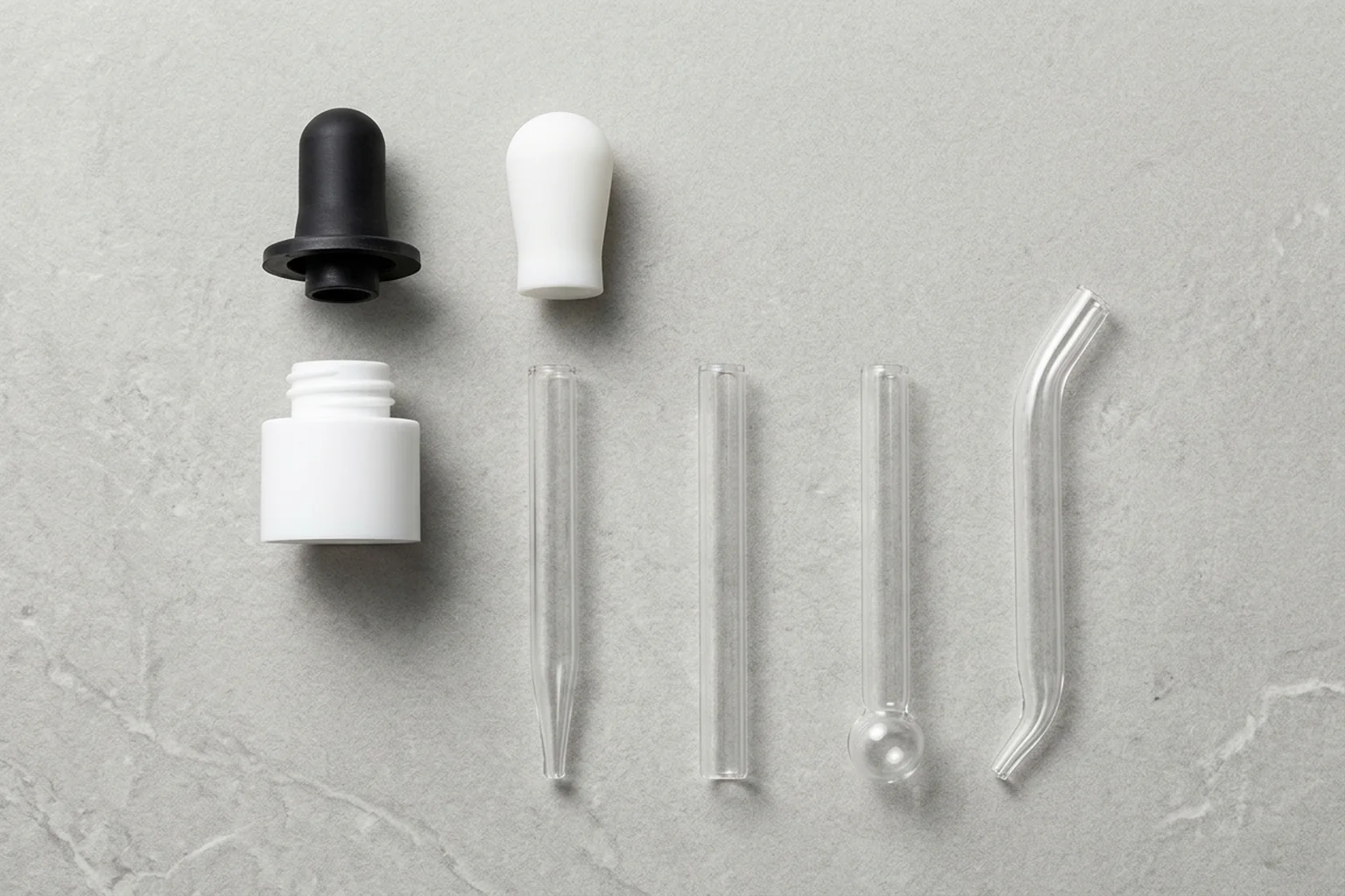 Overhead flat-lay arrangement of a black flanged rubber bulb, a white elastomer bulb, a white threaded collar cap, and four clear glass pipette tubes with straight, blunt, ball, and bent tips