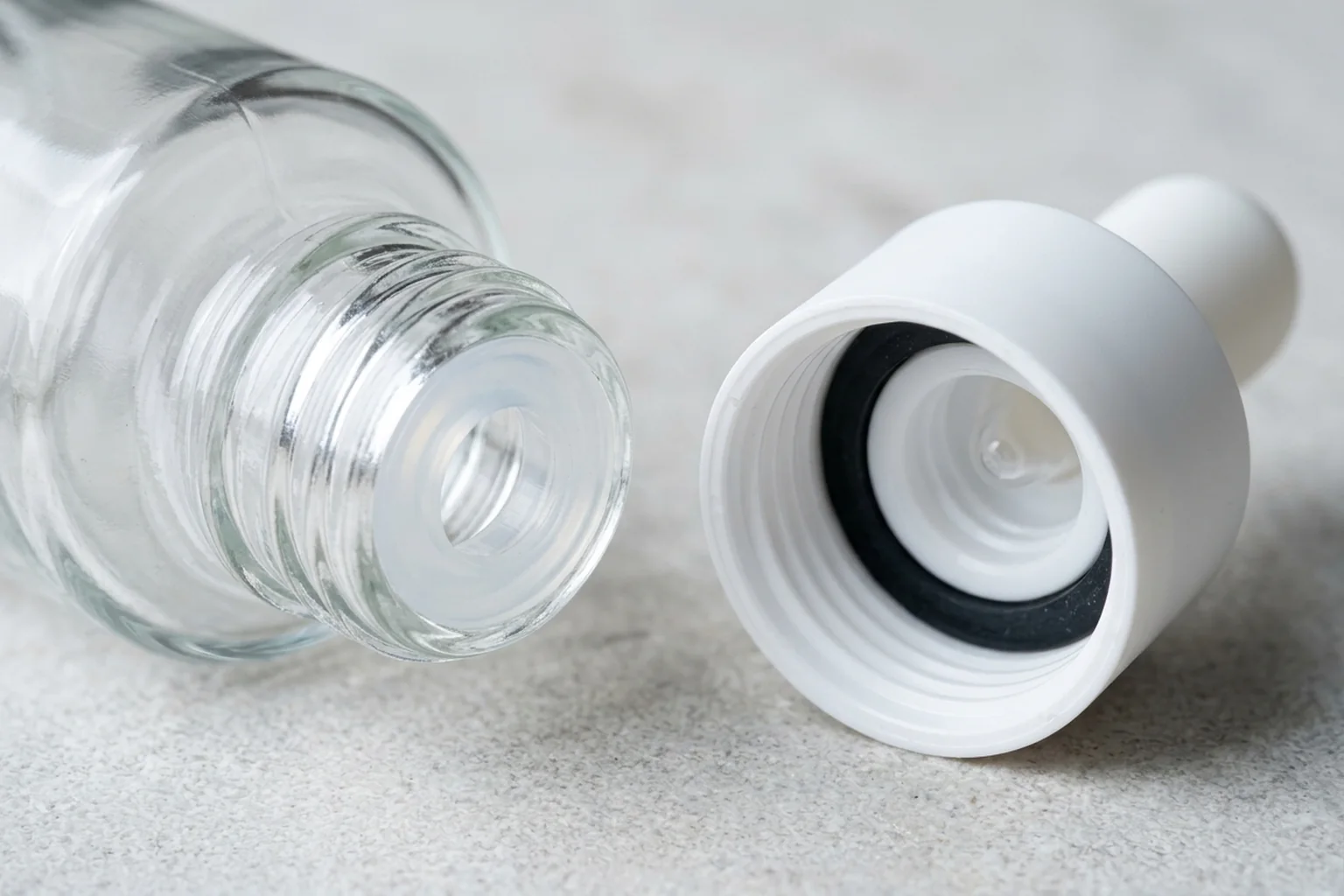 Macro close-up of a clear glass bottle neck fitted with a translucent internal wiper plug next to an upturned white dropper collar showing its black rubber underside flange gasket